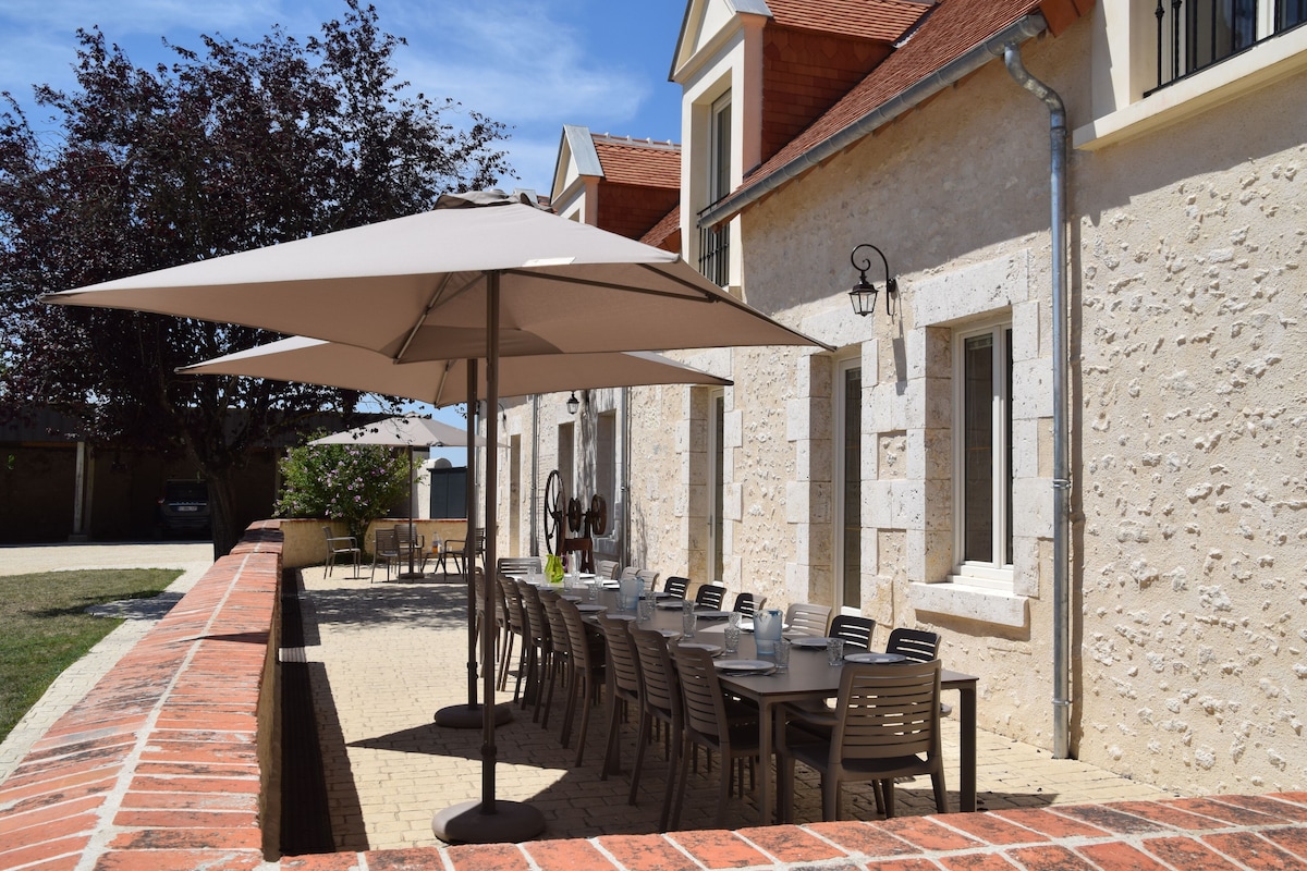 Une terrasse extérieure avec une grande table et des chaises confortables, abritée par un parasol, devant une maison en pierre avec des fenêtres et un mur en brique.