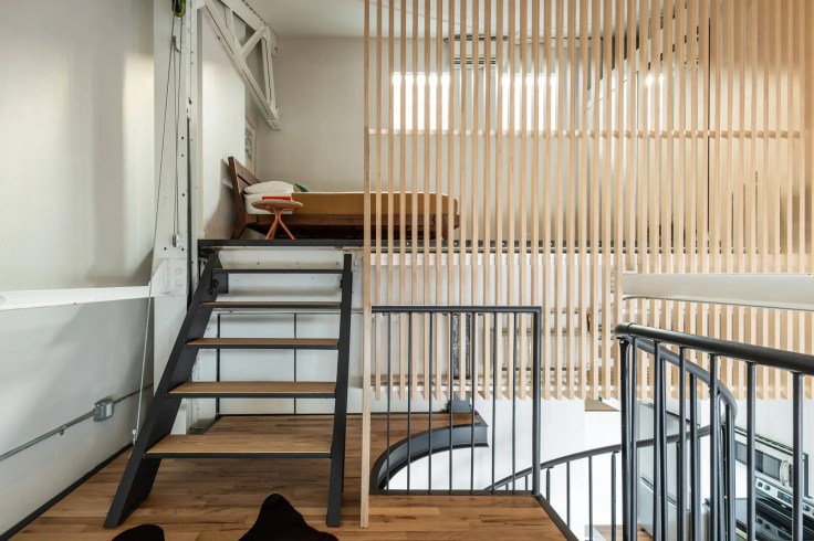 Vue intérieure d'un loft à deux étages avec un escalier menant à une mezzanine, séparée par des panneaux en bois, et une chambre au-dessus.