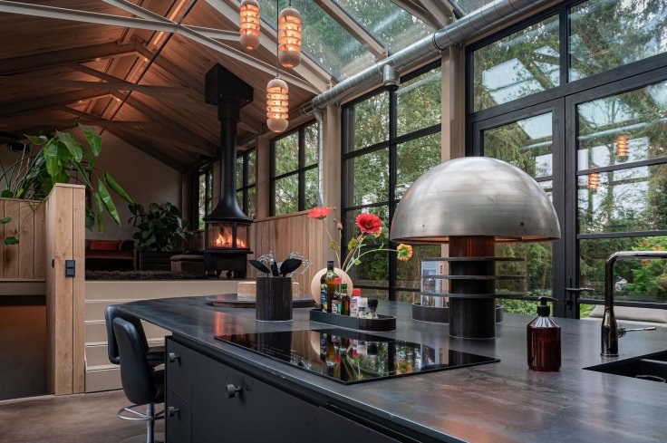 Intérieur d'une cuisine moderne avec îlot central, comprenant un évier et des appareils intégrés, entourée de baies vitrées offrant une vue sur la verdure environnante et un poêle à bois à l'arrière-plan.