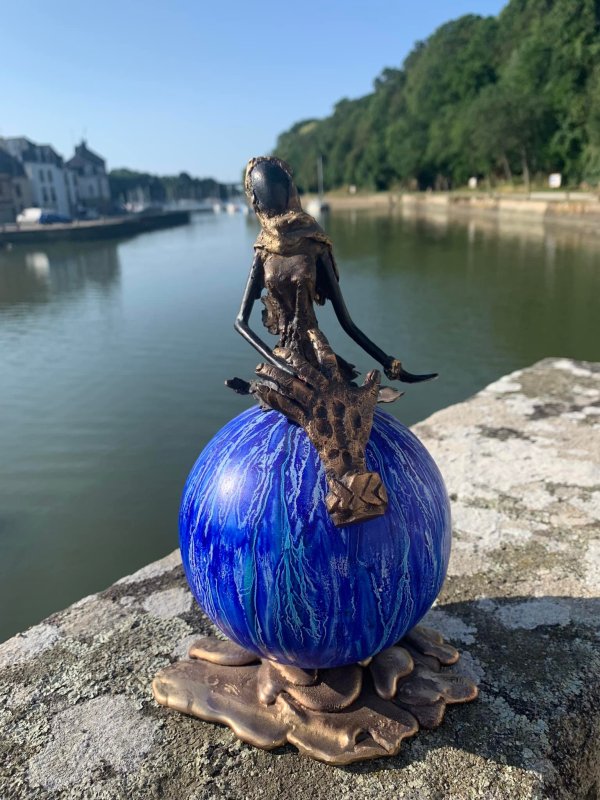A bronze sculpture of a seated figure on a blue spherical base, overlooking a serene waterway surrounded by greenery.