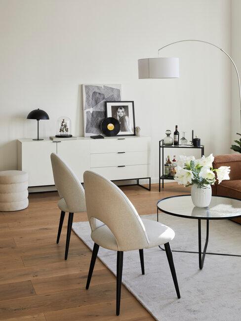 Interior of a modern living room featuring two beige chairs, a round glass coffee table, a side console with decorative items, and a cozy brown sofa. The space is well-lit with a floor lamp and includes various decor elements such as artwork and a vase of flowers.