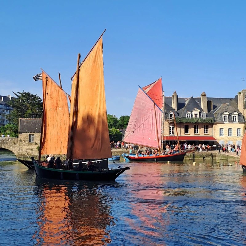 Trois voiliers avec des voiles orange et rouge naviguant sur l'eau calme du port de Saint-Goustan, avec des bâtiments historiques en arrière-plan et des personnes sur le quai.